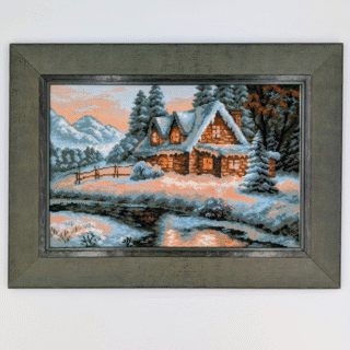 Framed cross-stitch artwork showing a snow-covered cabin with glowing windows beside a river, surrounded by pine trees and mountains at sunset.