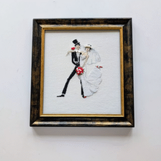 Framed cross-stitch artwork of a bride and groom dancing together in a stylized wedding tango, with black, white, and red accents.