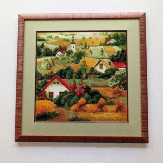 Framed cross-stitch artwork depicting a Serbian countryside with red-roofed houses, golden fields, trees, and rolling hills.