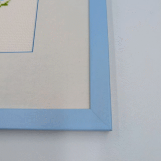 Framed cross-stitch artwork featuring a small bouquet of red poppies and bright blue cornflowers with green stems on a light background.