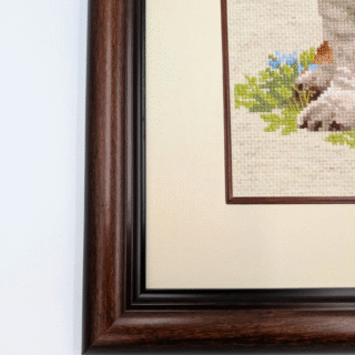 Framed cross-stitch artwork depicting a basset hound puppy sitting on grass with small blue flowers and tall meadow plants behind it.