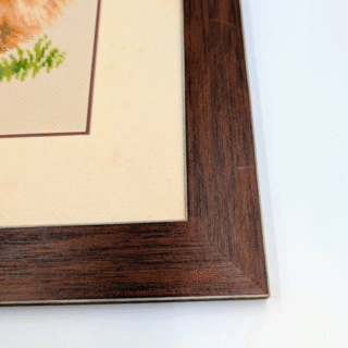 Framed cross-stitch artwork showing a fluffy Pomeranian dog standing in a garden with green foliage and a trellis in the background.