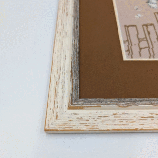 Framed cross-stitch artwork of a gray kitten perched on a tree stump above small flowers in a soft woodland scene.