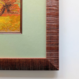 Framed cross-stitch artwork depicting a Serbian countryside with red-roofed houses, golden fields, trees, and rolling hills.