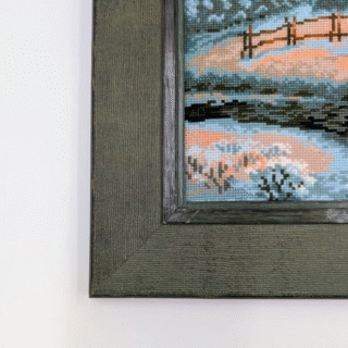 Framed cross-stitch artwork showing a snow-covered cabin with glowing windows beside a river, surrounded by pine trees and mountains at sunset.