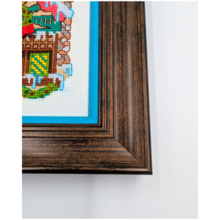 Stitched and framed embroidery artwork showing a festive gingerbread-style house decorated with wreaths, snow, and holiday details.