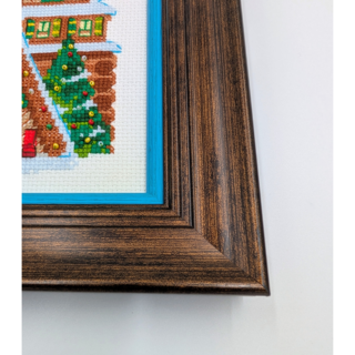 Stitched and framed embroidery artwork showing festive A-frame houses decorated with wreaths, trees, and winter details.