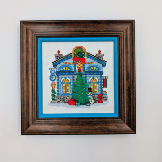 Stitched and framed embroidery artwork showing a festive townhouse with a decorated Christmas tree, gifts, and winter details.