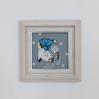 Framed and stitched cross-stitch artwork showing a small owl wearing a blue winter hat, ice skating among falling snowflakes on a soft gray background.
