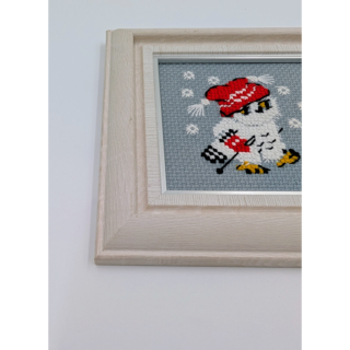 Stitched and framed embroidery artwork showing a small owl wearing a red winter hat against a soft gray background with snowflakes.