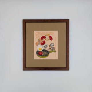 Stitched and framed embroidery artwork depicting a playful breakfast scene with a fried egg, tomatoes, bread, coffee items, and a skillet arranged in mid-air.