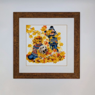 Stitched and framed embroidery artwork showing two children playing with a dog in a pile of golden autumn leaves.