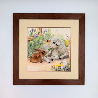 Framed stitched artwork depicting a lamb and a small rabbit resting together among spring flowers in a peaceful garden scene.