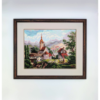 Stitched and framed embroidery artwork depicting an alpine village with a church, hillside homes, and mountains in the background.