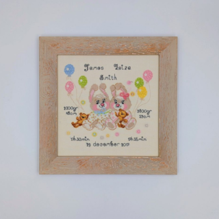 Stitched and framed embroidery artwork featuring two baby bunnies with balloons, teddy bears, and personalized birth details on a soft pastel background.