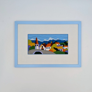 Framed stitched artwork showing a colorful village with small houses, a church tower, and rolling hills beneath a blue sky.