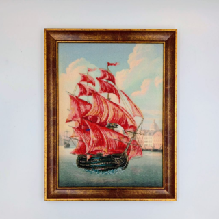 Framed stitched artwork showing a sailing ship with vivid red sails gliding across calm water against a soft pastel sky.
