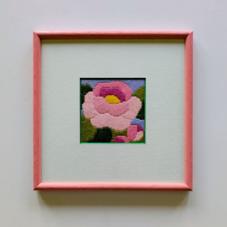 Framed stitched artwork featuring a stylized pink flower with layered petals and green foliage on a soft pastel background.