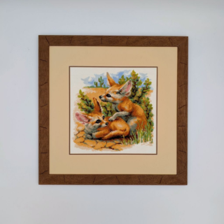 Framed stitched artwork depicting two desert foxes resting among rocks and shrubs in a warm, natural landscape.