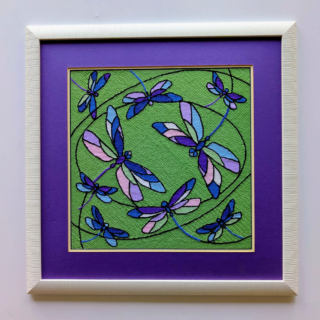 Stitched and framed embroidery artwork featuring colorful dragonflies flying across a green background with flowing lines.