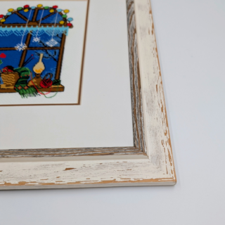 Framed stitched artwork showing a cozy winter window scene with potted plants, yarn, candles, and soft lights against a deep blue evening sky.