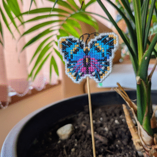Small cross-stitched butterfly decoration on a stick, placed among green leaves.