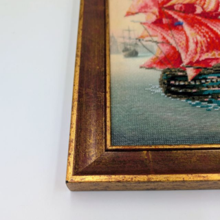 Framed stitched artwork showing a sailing ship with vivid red sails gliding across calm water against a soft pastel sky.