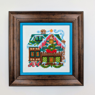 Stitched and framed embroidery artwork showing a festive gingerbread-style house decorated with wreaths, snow, and holiday details.