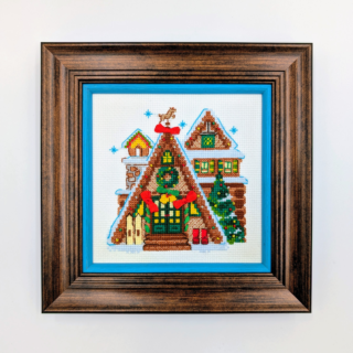 Stitched and framed embroidery artwork showing festive A-frame houses decorated with wreaths, trees, and winter details.