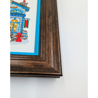Stitched and framed embroidery artwork showing a festive townhouse with a decorated Christmas tree, gifts, and winter details.