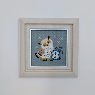 Stitched and framed embroidery artwork showing a small owl beside an alarm clock on a soft gray background.