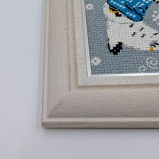 Framed and stitched cross-stitch artwork showing a small owl wearing a blue winter hat, ice skating among falling snowflakes on a soft gray background.