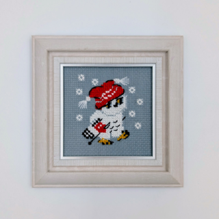 Stitched and framed embroidery artwork showing a small owl wearing a red winter hat against a soft gray background with snowflakes.