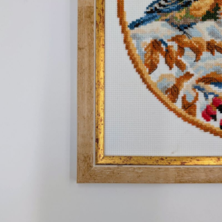 Stitched and framed embroidery artwork showing a small bird perched in a snowy winter landscape framed by autumn leaves in a circular composition.