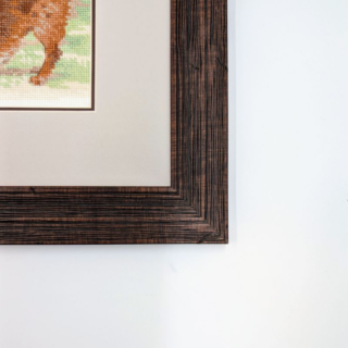 Stitched and framed embroidery artwork showing a young boy holding a football while two playful dogs run and splash nearby on a grassy field.
