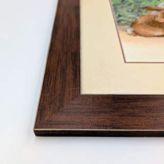 Framed stitched artwork depicting a lamb and a small rabbit resting together among spring flowers in a peaceful garden scene.