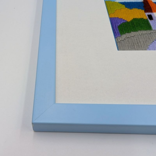 Framed stitched artwork showing a colorful village with small houses, a church tower, and rolling hills beneath a blue sky.