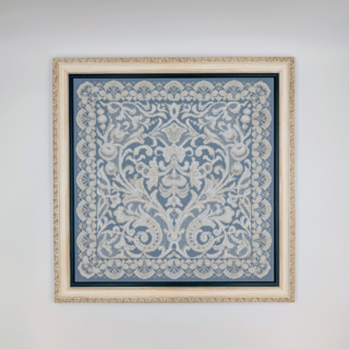 Framed stitched artwork featuring an intricate lace-style ornamental pattern in soft ivory tones on a muted blue background.