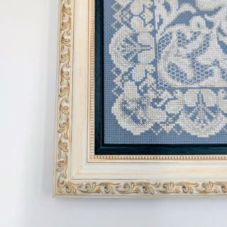 Framed stitched artwork featuring an intricate lace-style ornamental pattern in soft ivory tones on a muted blue background.
