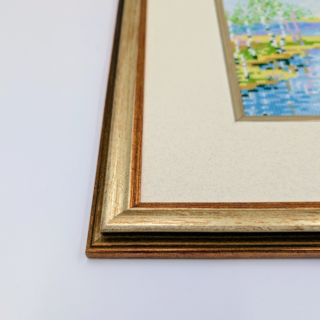 Framed stitched artwork depicting a calm river flowing between grassy banks and slender trees under a soft, clouded sky.