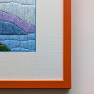 Framed stitched artwork showing a colorful coastal village with small houses, trees, hills, and water in soft blue and pastel tones.