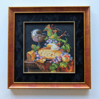 Stitched and framed embroidery artwork depicting a still life with cheese, grapes, fruit, and a wine goblet on a dark background.