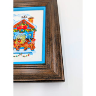 Stitched and framed embroidery artwork showing a festive winter village scene with decorated houses and a horse-drawn sleigh.