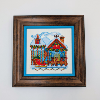 Stitched and framed embroidery artwork showing a festive winter village scene with decorated houses and a horse-drawn sleigh.