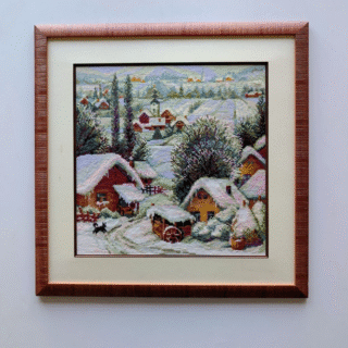 Framed cross-stitched artwork depicting a snowy Serbian village with small wooden houses, trees, and winding paths set among rolling winter hills.