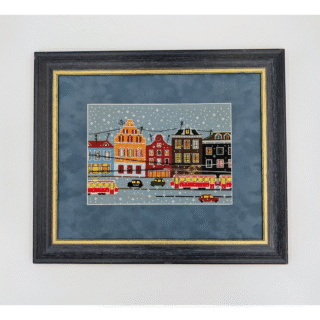 Framed cross-stitch artwork showing colorful city buildings, trams, and cars on a snowy winter day.