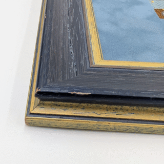 Framed cross-stitch artwork showing a colorful city street in autumn with falling leaves, bicycles, and cars.