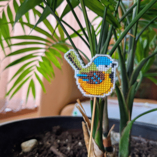 Small cross-stitched bird decoration on a stick, displayed among green leaves.