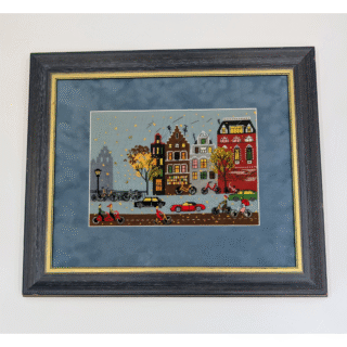 Framed cross-stitch artwork showing a colorful city street in autumn with falling leaves, bicycles, and cars.