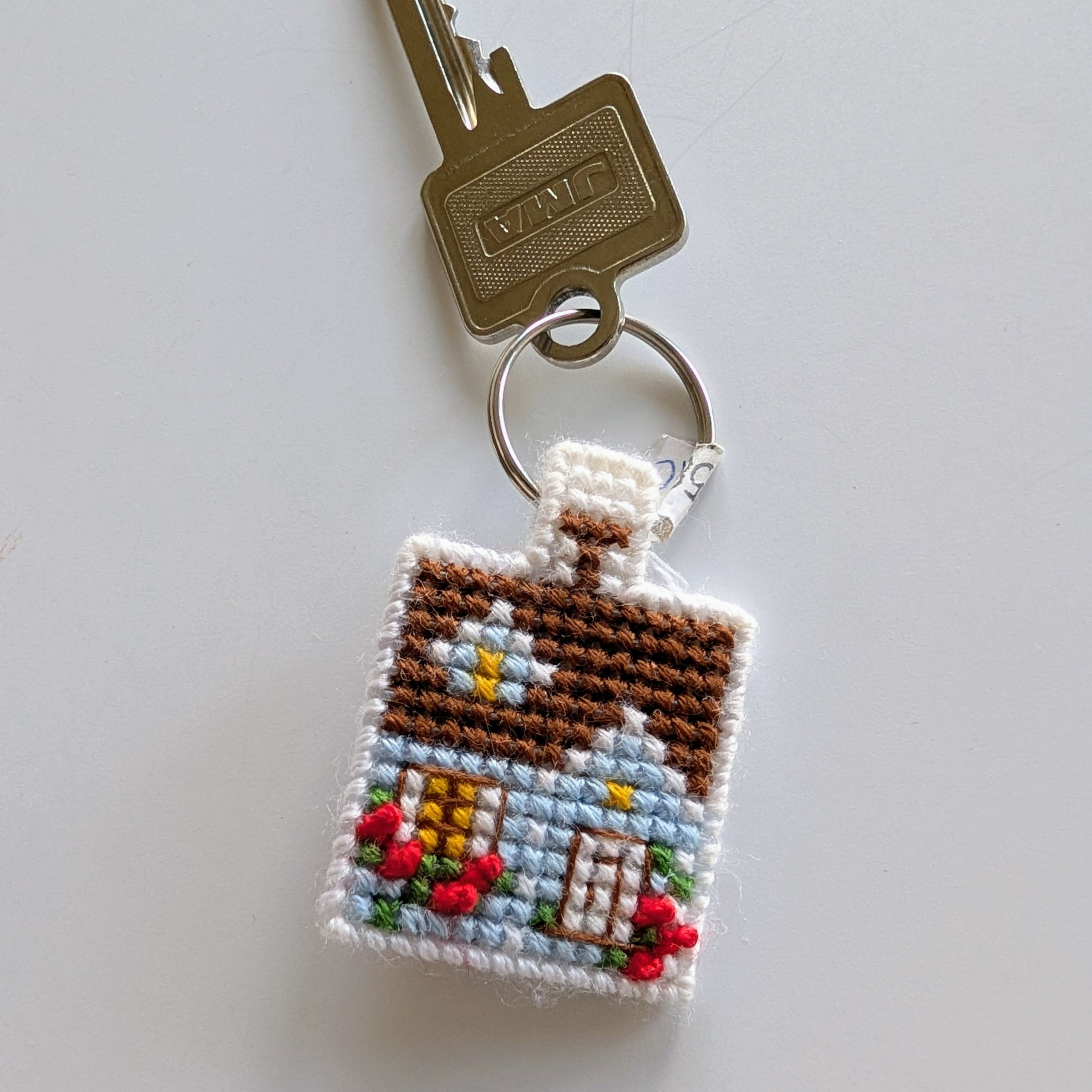 Small cross-stitched keychain featuring a tiny blue house with a brown roof and red flowers.